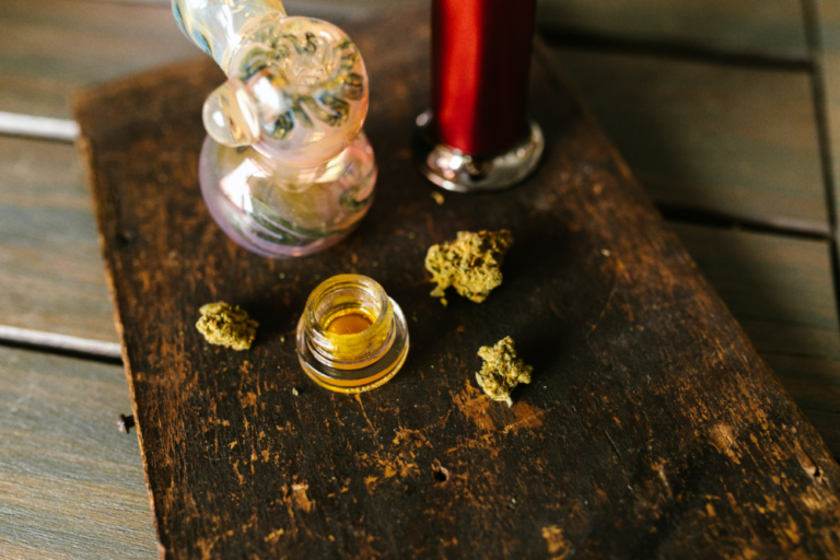 A close-up photo of a glass jar containing cannabis concentrate, with a cannabis plant and dispensary products in the background.